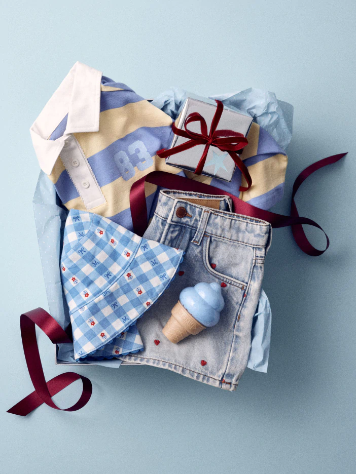 Flat lay of summer clothing and accessories on light blue background: striped shirt, denim shorts, gingham fabric, ice cream toy, and burgundy ribbon