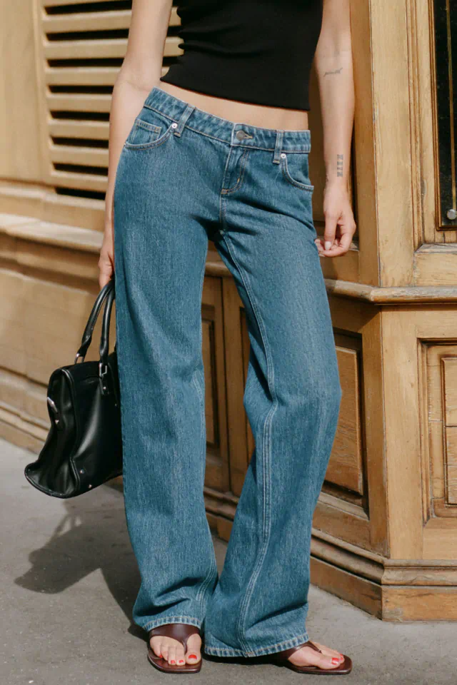 Woman wearing low-rise blue jeans and black crop top, holding a black handbag, standing against wooden doors
