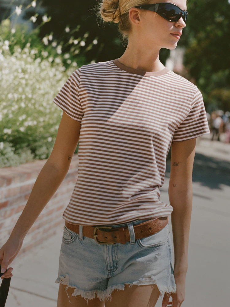 Woman wearing brown and white striped short-sleeve t-shirt and distressed denim shorts with brown belt, standing outdoors wearing sunglasses