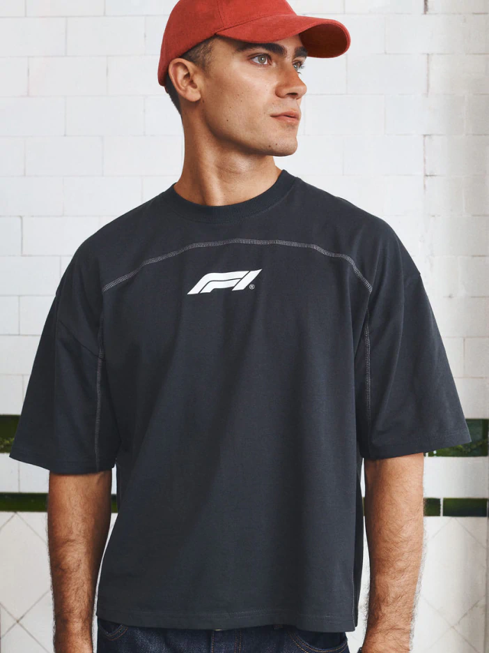 Man wearing black F1 logo t-shirt and red cap, standing against white brick wall
