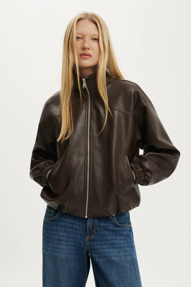 Woman with long blonde hair wearing a black leather zip-up jacket over a white top and blue jeans against a white background