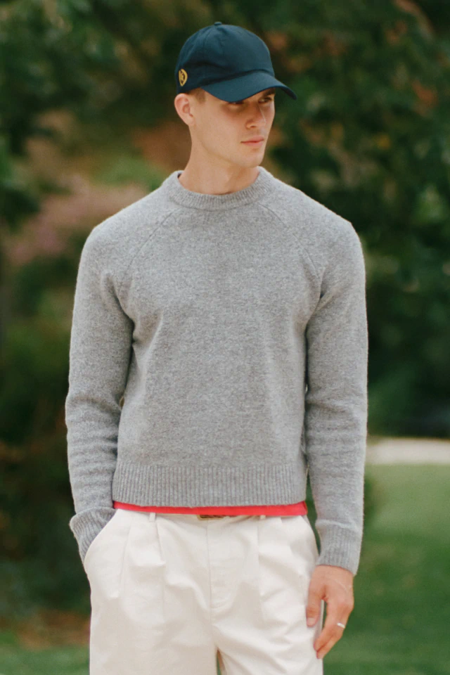 Male model wearing gray crew neck sweater with red waistband detail and cream pants, navy cap, outdoors