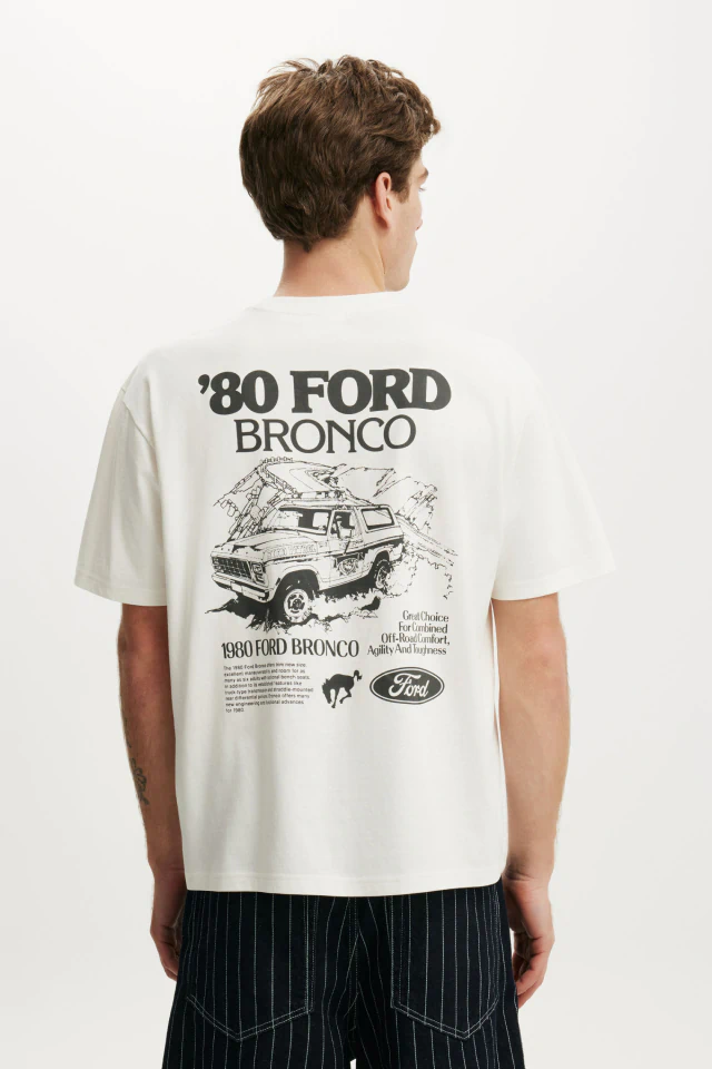 Man wearing cream '80 Ford Bronco' graphic t-shirt with vehicle illustration, paired with black pinstriped pants, shown from behind