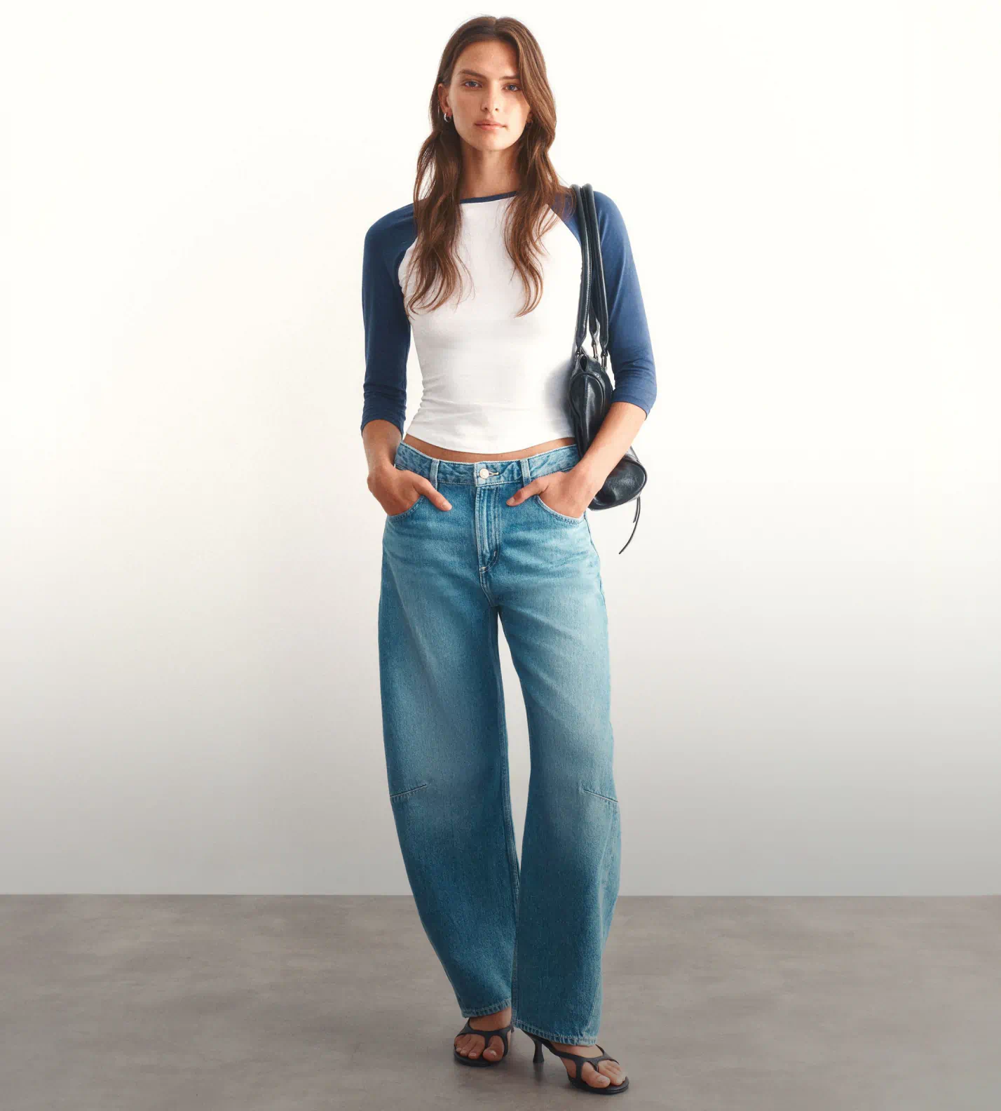 Woman wearing navy blue three-quarter sleeve raglan shirt over white cropped tee, blue denim jeans, and black slide sandals, carrying a black shoulder bag