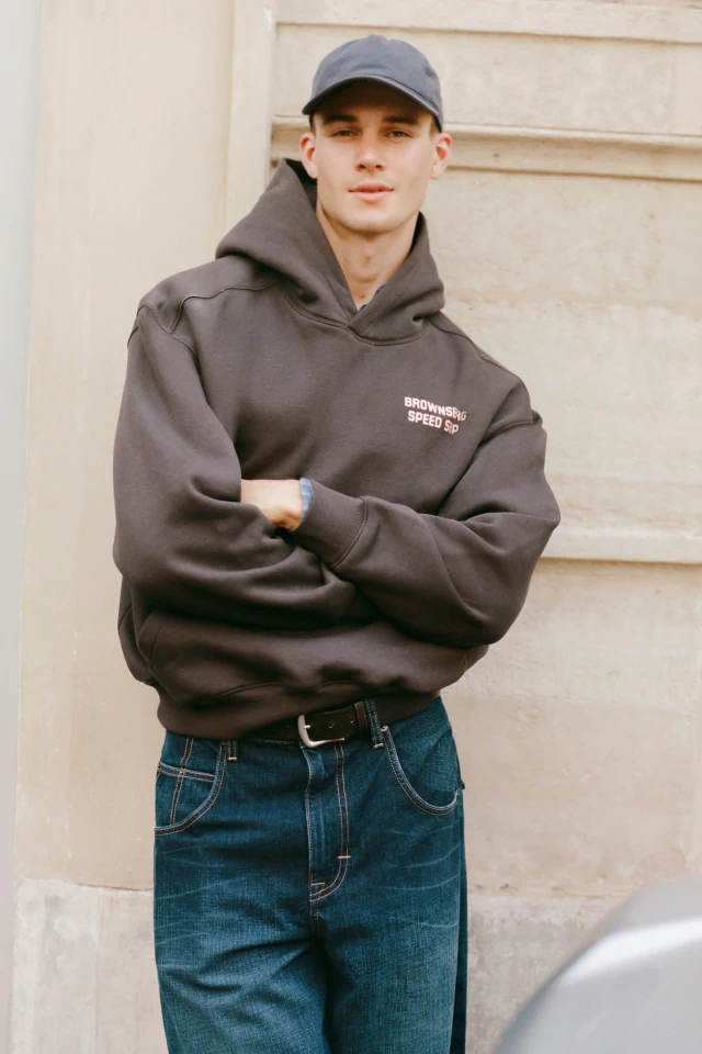 Young man wearing charcoal hoodie with "Drowning Speed Shop" text, blue cap, and dark jeans against neutral background