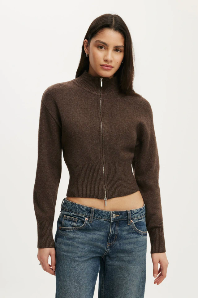 Woman wearing a brown cropped zip-up cardigan with ribbed details and blue denim jeans against a white background