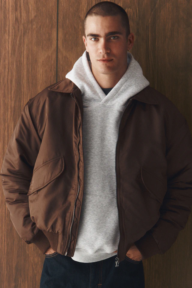 Man wearing brown zip-up jacket over gray hoodie and dark jeans, standing against wooden background