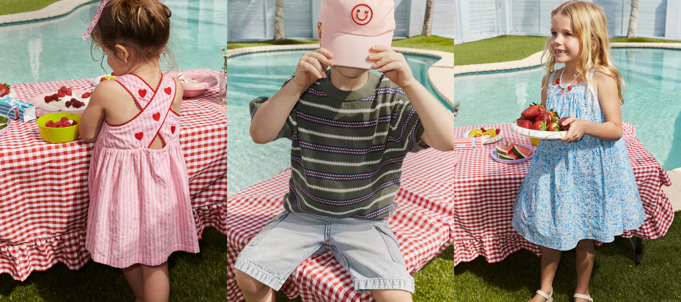 Three children at a poolside picnic table with red and white checkered tablecloth, holding food and wearing colorful summer dresses and sweaters