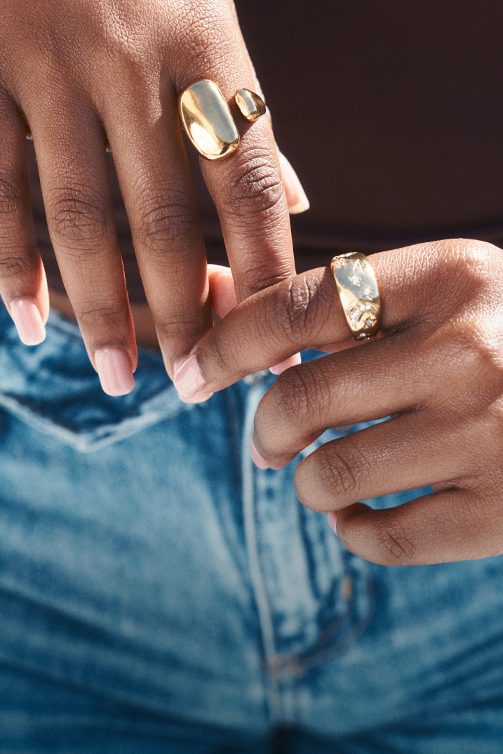 Close-up of hands wearing gold rings and pale pink nail polish against a blue denim background