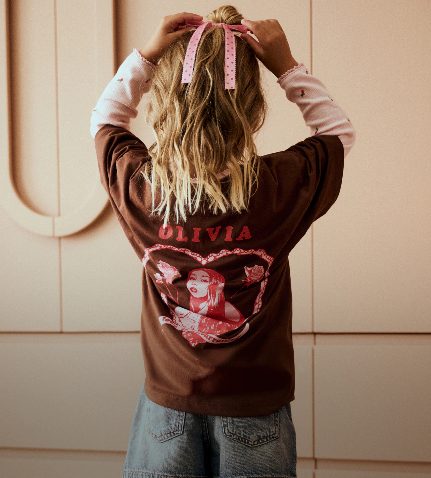 Back view of person wearing brown oversized shirt with "OLIVIA" text and red graphic print, styled with pink ribbon hair accessory and light blue jeans