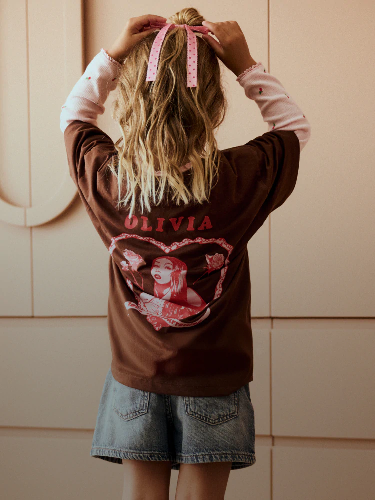 Person from behind wearing brown graphic sweatshirt with "OLIVIA" text and pink ribbon in long blonde hair, denim skirt