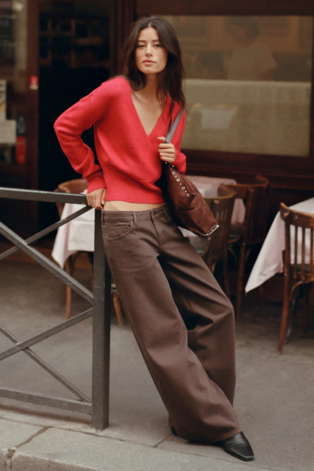 Woman wearing a bright red cropped V-neck sweater and brown wide-leg jeans, holding a brown shoulder bag on a city street