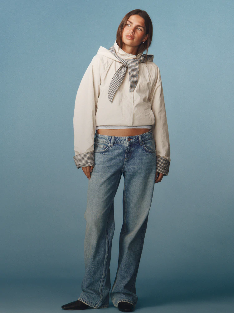 Woman wearing navy and white raglan top, light-wash wide-leg jeans, black belt, and black shoulder bag against blue background