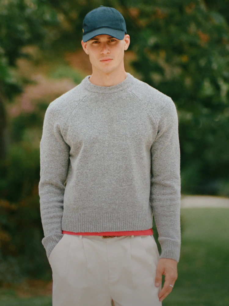 Man wearing gray crew neck sweater with red trim and beige pants, blue cap, standing outdoors against blurred green background