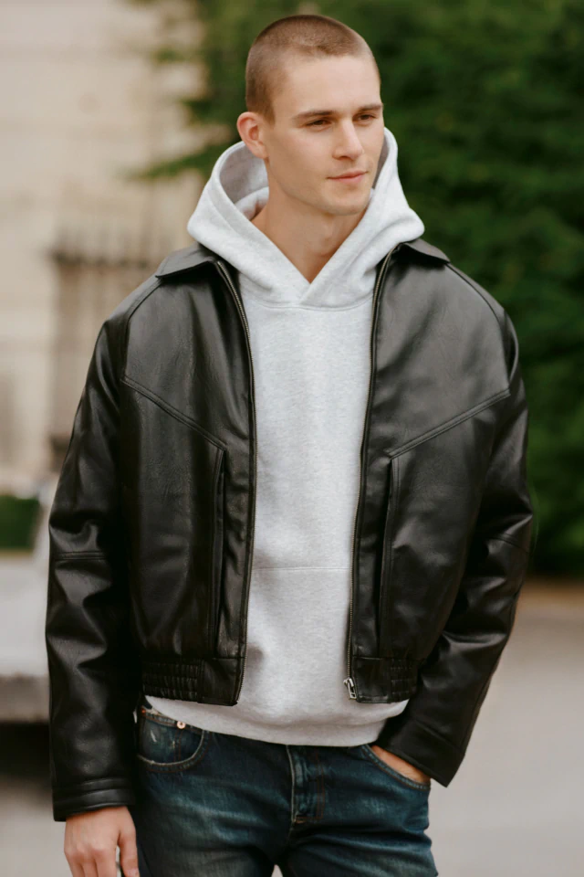 Young man wearing black leather jacket over light gray hoodie and dark blue jeans, standing outdoors against blurred green foliage