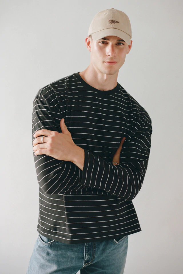 Man wearing cream baseball cap and black striped long-sleeve t-shirt with blue jeans, posed against neutral background