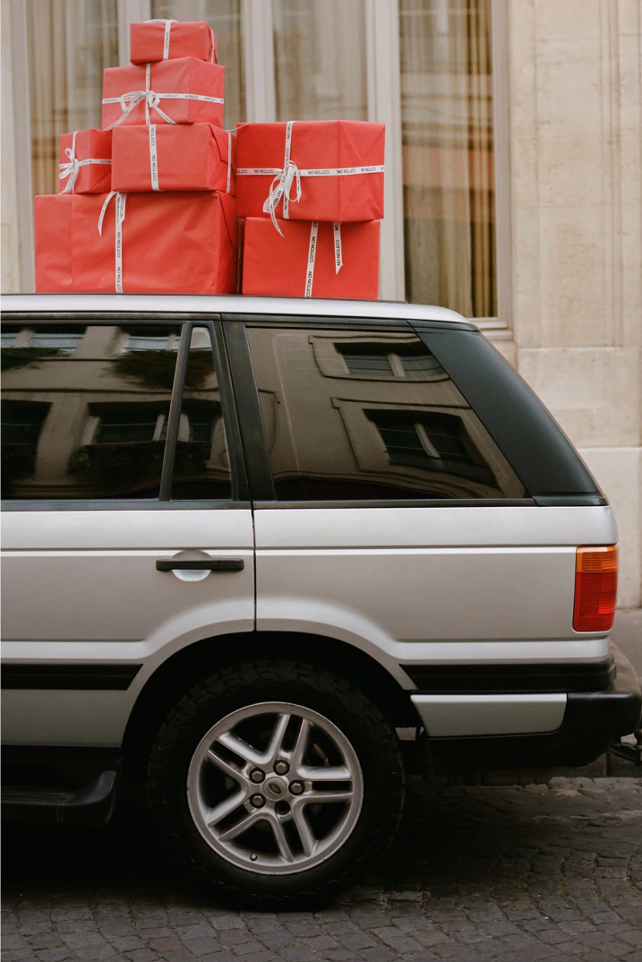 Silver SUV loaded with red gift boxes tied with white ribbons on roof, parked in front of elegant building with cream curtains