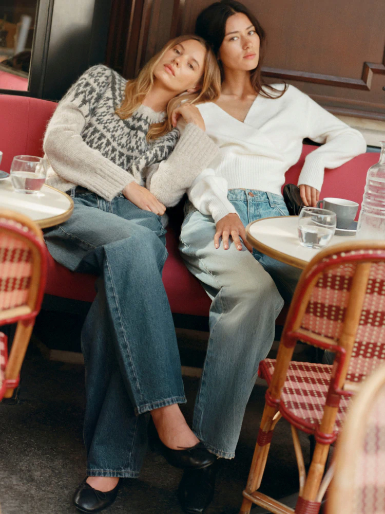 Two women seated at a restaurant table wearing knit sweaters and jeans, looking upward thoughtfully