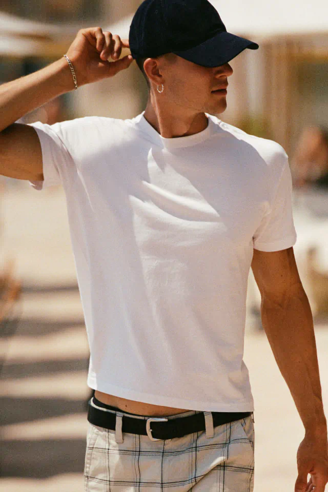 Man wearing white crew neck t-shirt, black baseball cap, and plaid shorts with black belt, adjusting cap outdoors