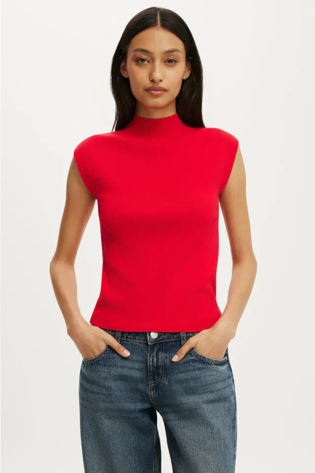Woman wearing a red sleeveless mock-neck top and blue jeans against a white background