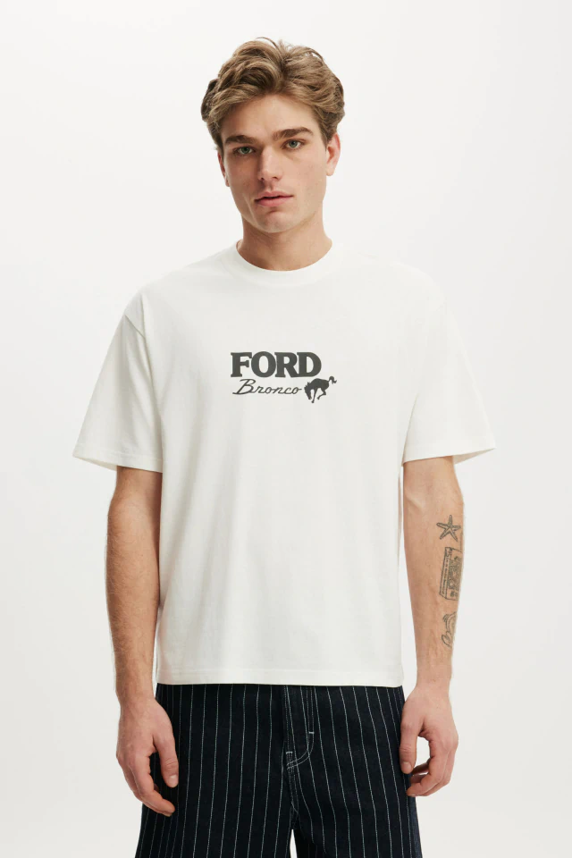 Man wearing cream Ford Bronco graphic t-shirt and black pinstriped pants against white background