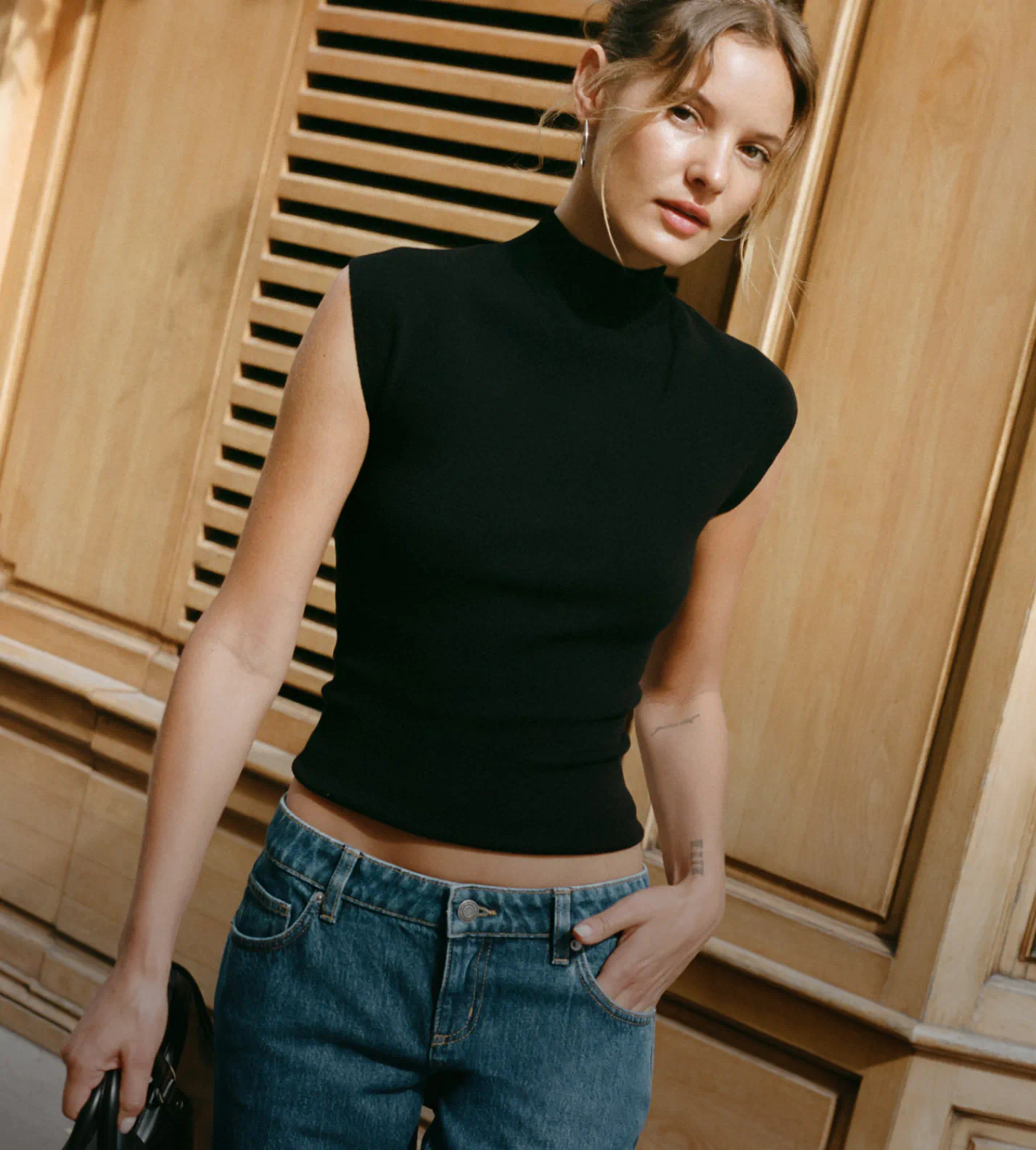 Woman wearing black sleeveless turtleneck and blue jeans against wooden paneled background