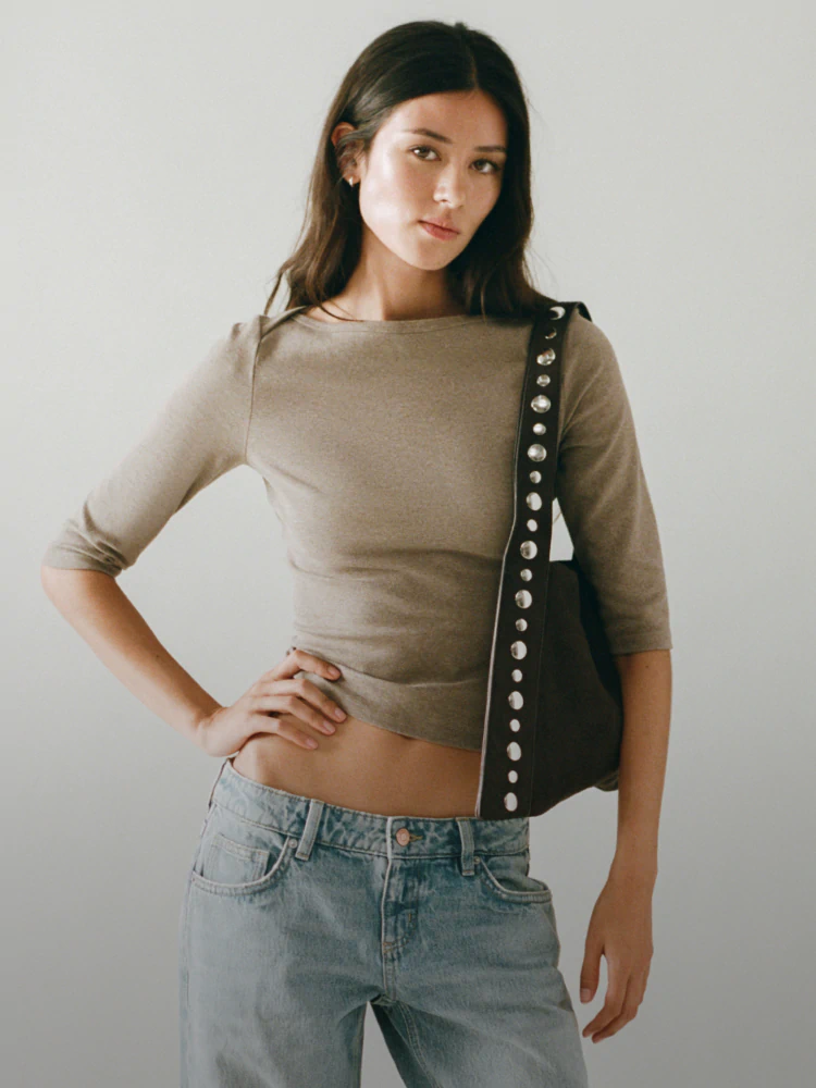 Woman wearing beige three-quarter sleeve crop top and light blue jeans with black studded shoulder bag against white background