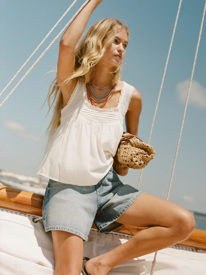Woman wearing white sleeveless tank top and denim shorts on a sailboat, holding bread, against blue sky