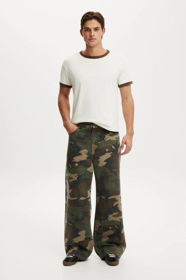 Man wearing cream ringer t-shirt and camouflage wide-leg jeans standing against white background