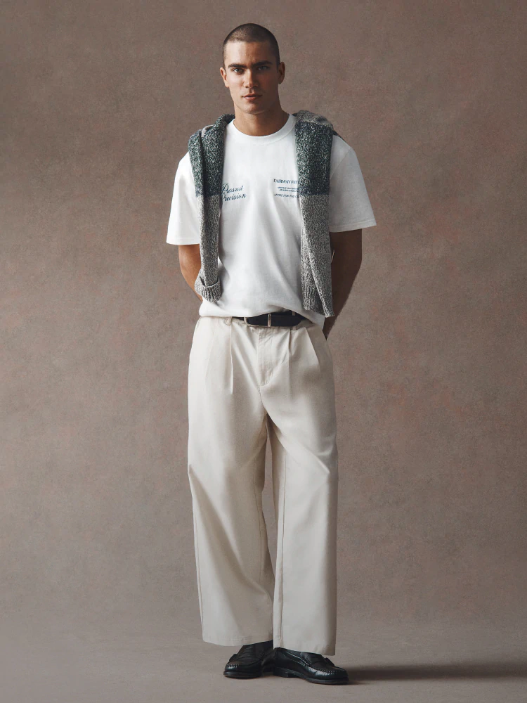 Man wearing white graphic t-shirt, cream wide-leg trousers, gray knit vest, and black leather shoes against neutral background