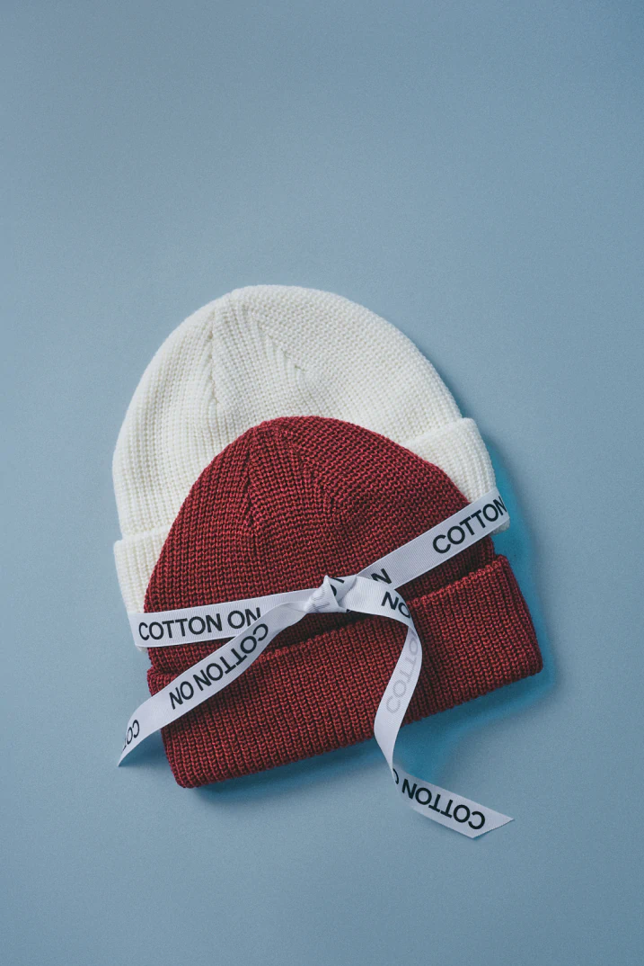 Two knit beanies stacked together—white on top, burgundy below—wrapped with white "COTTON ON" branded ribbon against a blue background