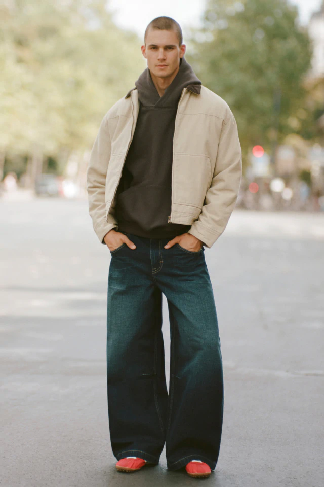 Male model wearing beige jacket over charcoal hoodie, dark blue jeans, and red sneakers, standing outdoors