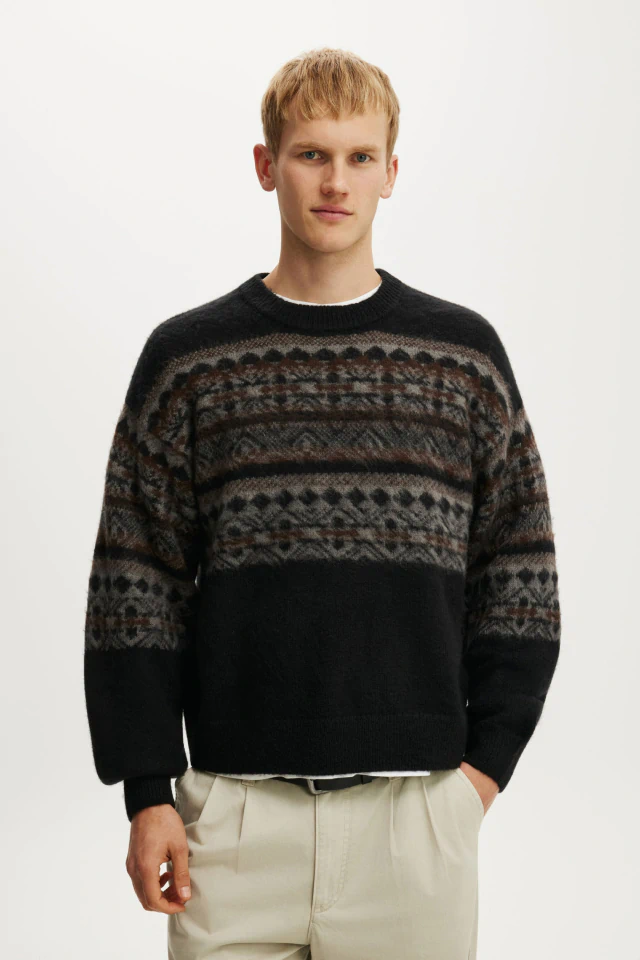 Male model wearing black crew neck sweater with brown and grey patterned stripes, paired with cream trousers against white background