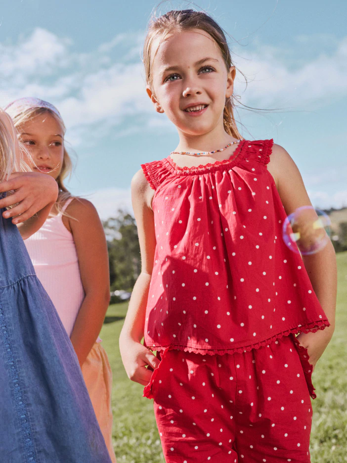Young girl wearing red polka dot sleeveless top and matching pants standing outdoors with friends on a sunny day