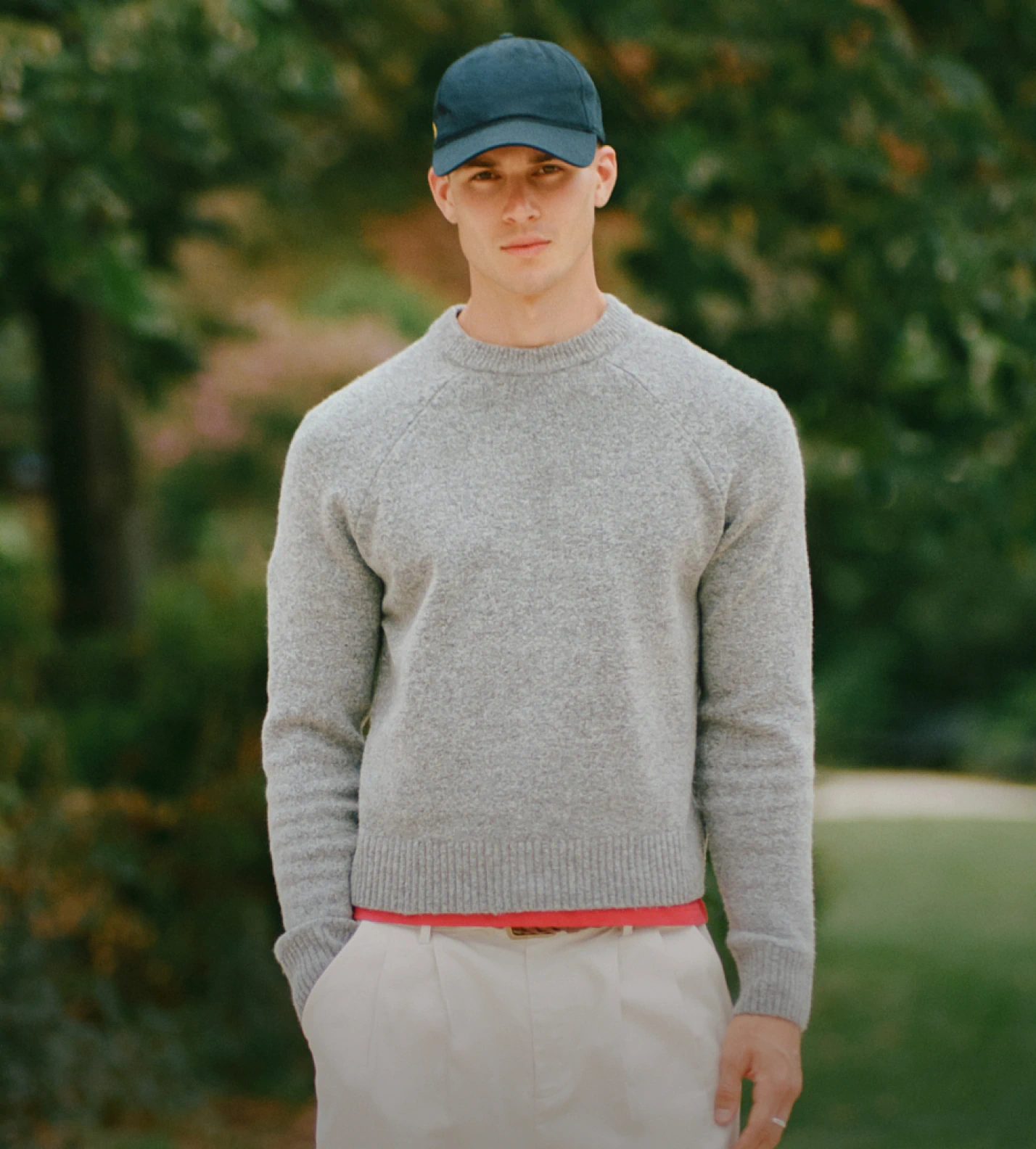Man wearing light gray crew neck sweater with red waistband detail and cream pants, navy cap, standing outdoors against blurred green foliage