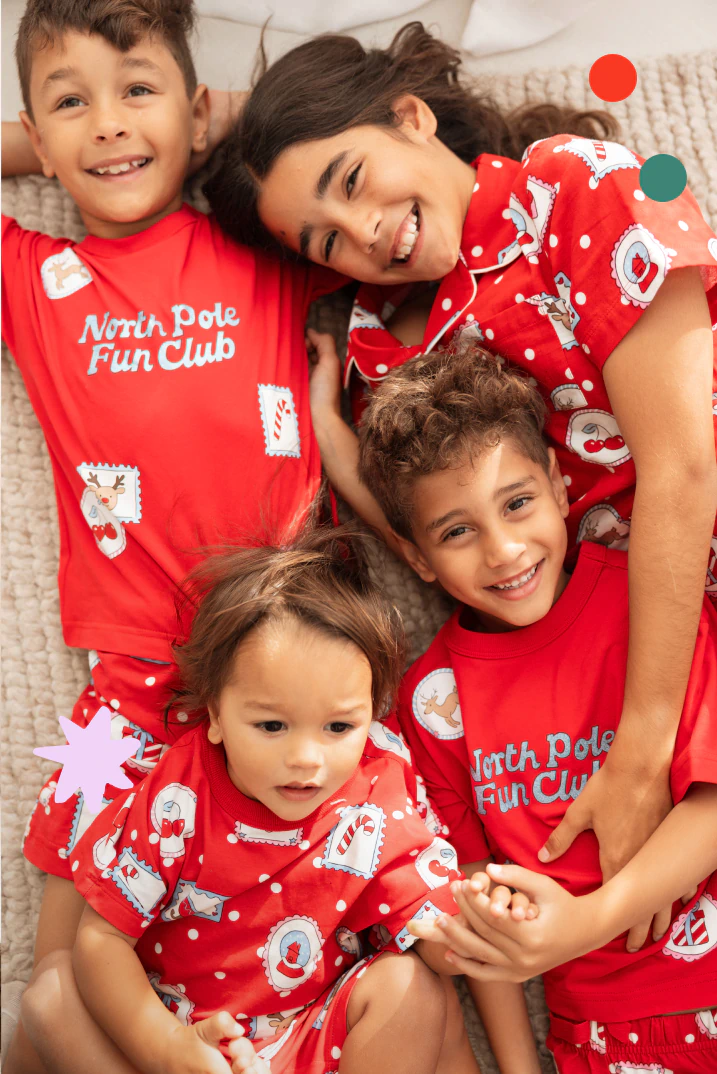 Family of four wearing matching red "North Pole Fun Club" Christmas pajamas with festive prints, smiling together on a couch