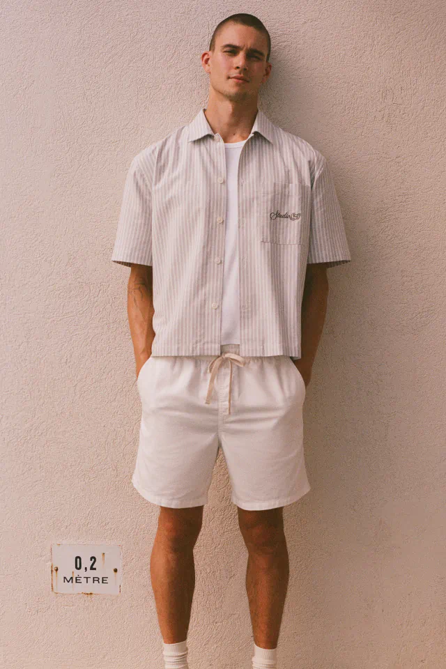 Man wearing light pink striped short-sleeve shirt over white t-shirt and cream drawstring shorts against beige wall