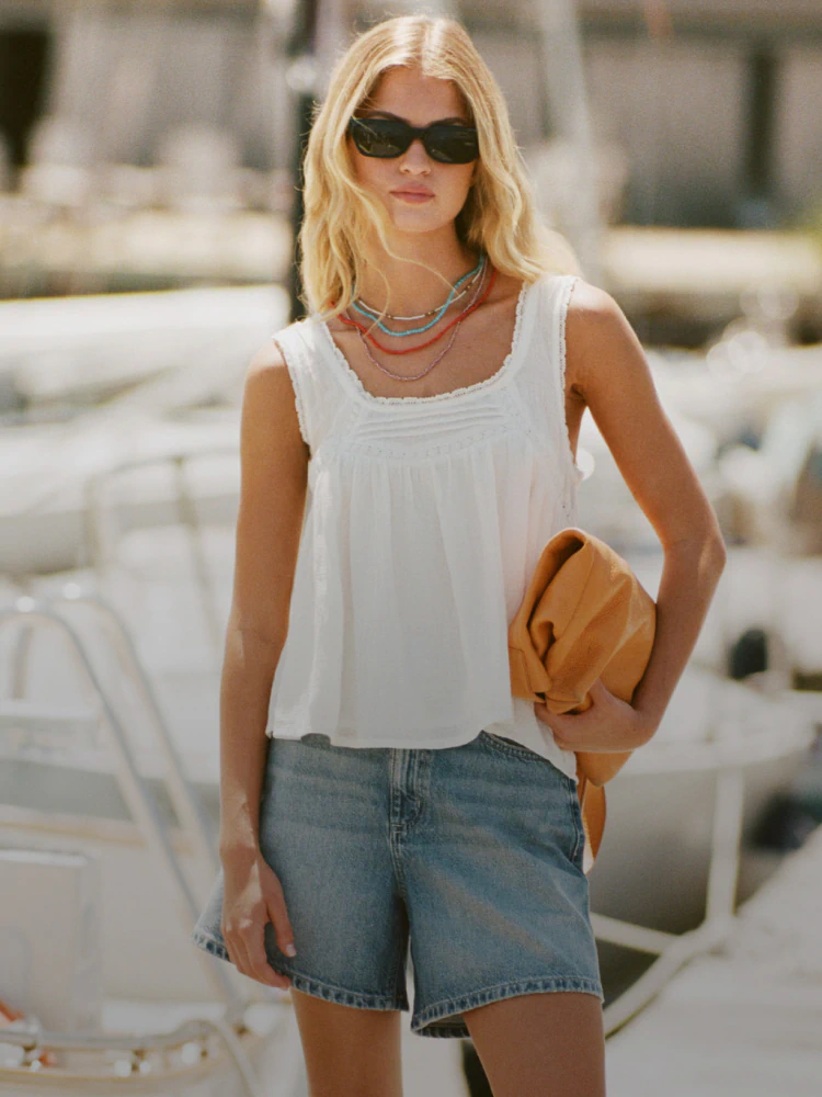 Woman wearing white sleeveless tank top and denim shorts with brown shoulder bag and dark sunglasses, standing outdoors