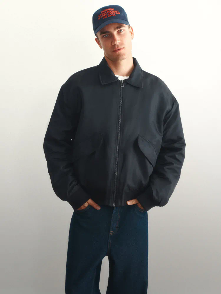 Man wearing navy zip-up jacket over white shirt and dark blue jeans, styled with navy cap against cream background