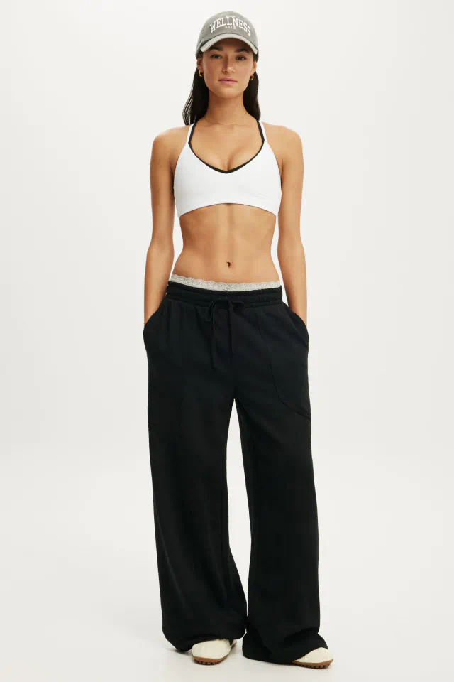 Woman wearing white sports bra with black trim, black wide-leg lounge pants, and gray "Wellness" baseball cap against white background