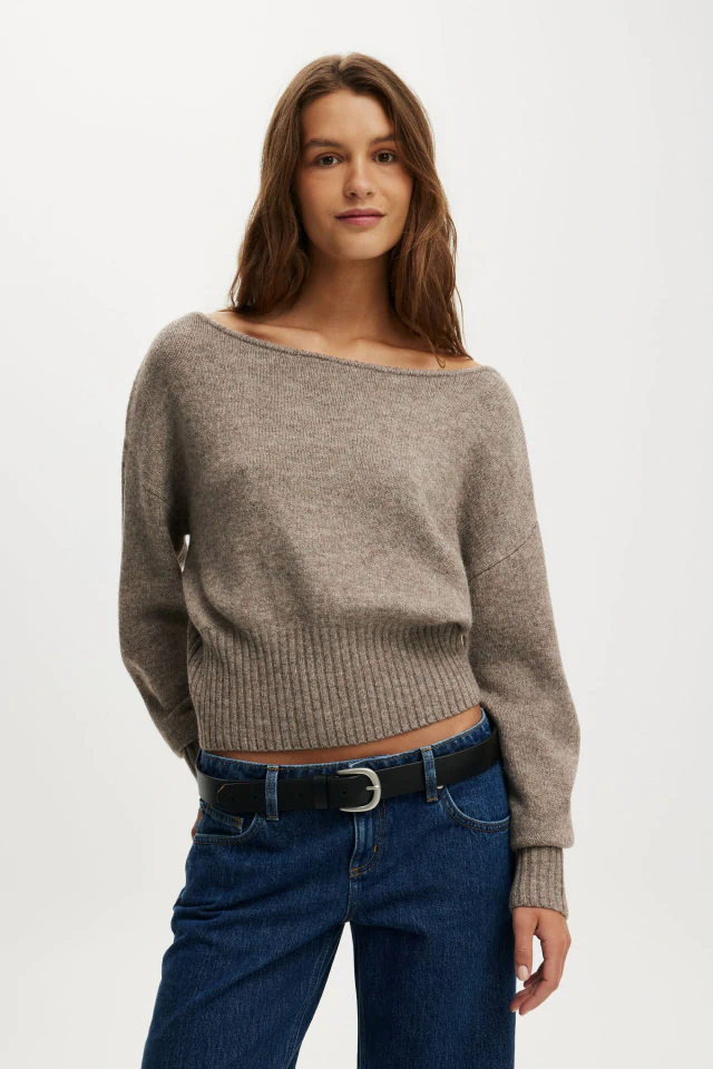Woman wearing a beige ribbed cropped sweater with long sleeves and dark blue jeans with a black belt against a white background