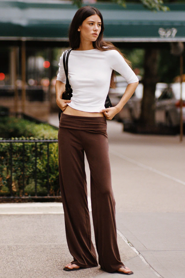 Woman wearing white cropped half-sleeve top and brown wide-leg pants, carrying black shoulder bag, standing on urban street