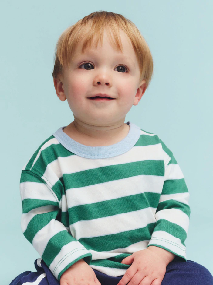 Toddler with reddish-blonde hair wearing a green and white striped long-sleeve sweater and navy pants against a light blue background