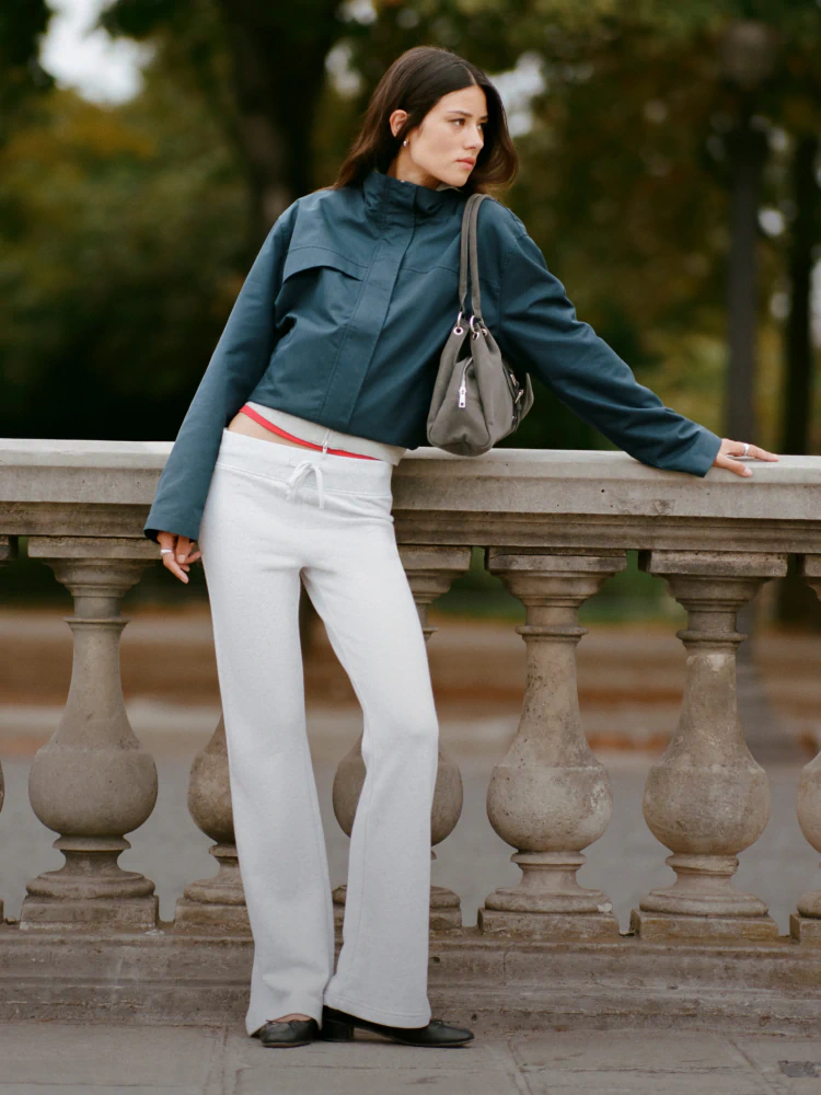 Woman wearing teal cropped jacket and white wide-leg pants with red waistband detail, holding grey shoulder bag, posed against stone balustrade