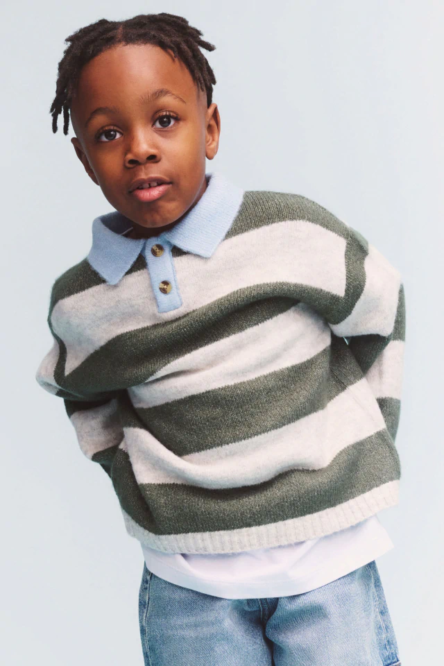 Young child wearing a beige zip-up jacket over a dark and white striped shirt and blue jeans, smiling at camera