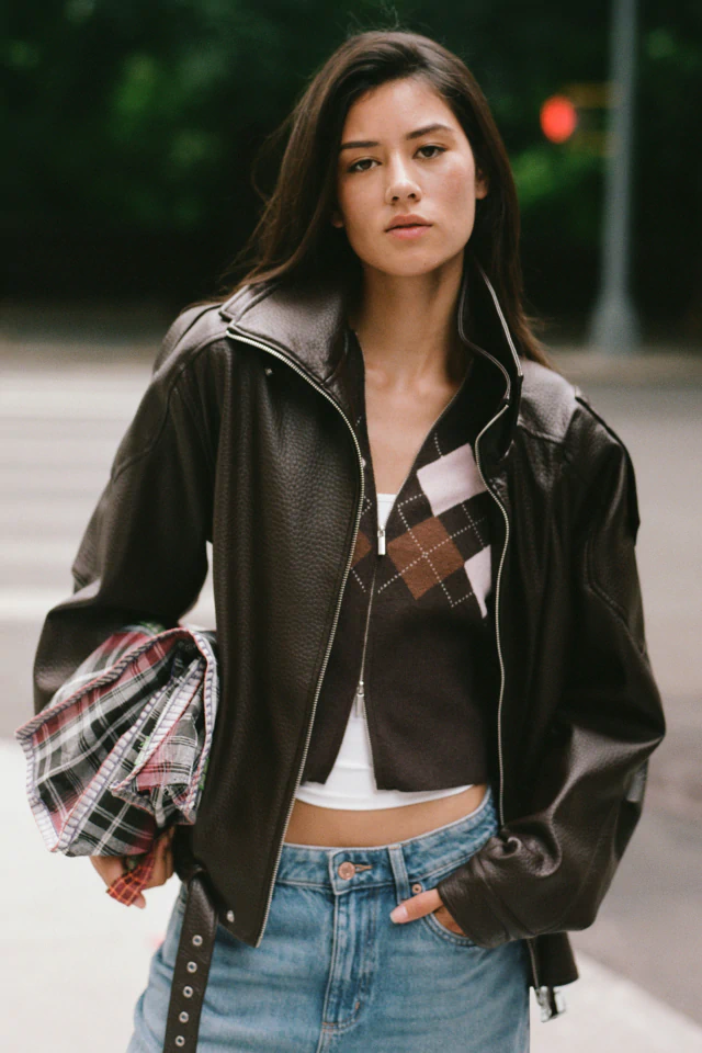 Woman wearing a black leather jacket over an argyle sweater and white crop top, paired with blue jeans, holding a plaid shirt