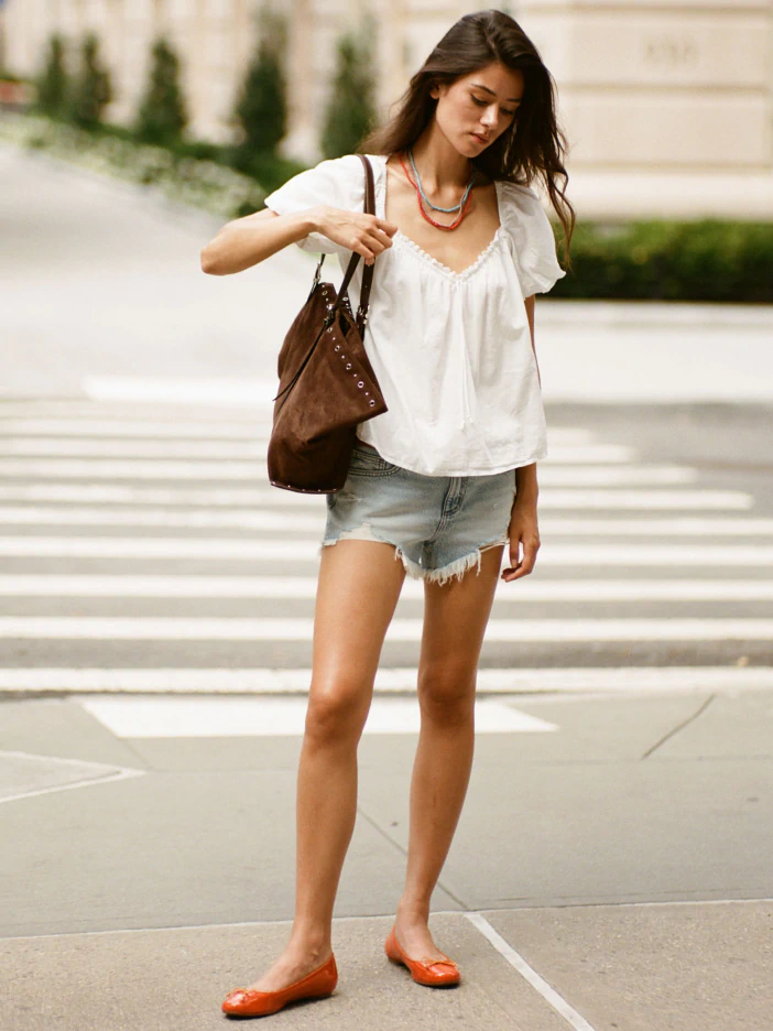 Woman wearing white v-neck blouse and light denim shorts with brown shoulder bag and orange ballet flats, standing on urban street