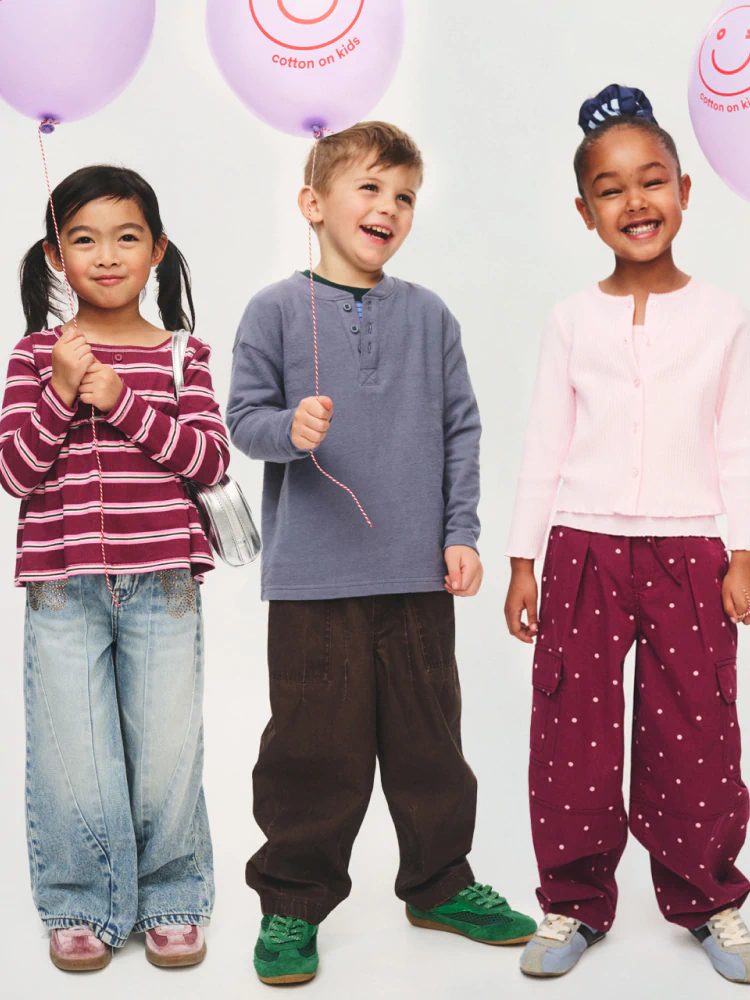 Three smiling children holding purple balloons printed with "Cotton on Kids" logo, wearing colorful casual clothing against white background