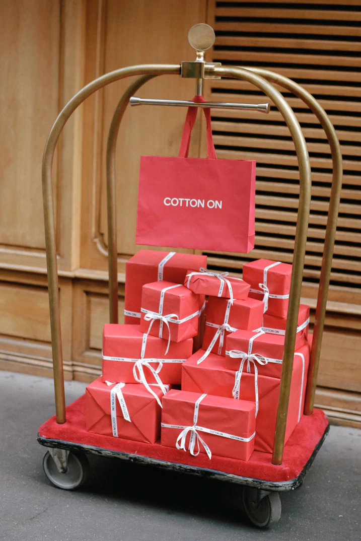 Cotton On branded bellhop cart display with red gift boxes tied with white ribbon and a red shopping bag against wooden backdrop