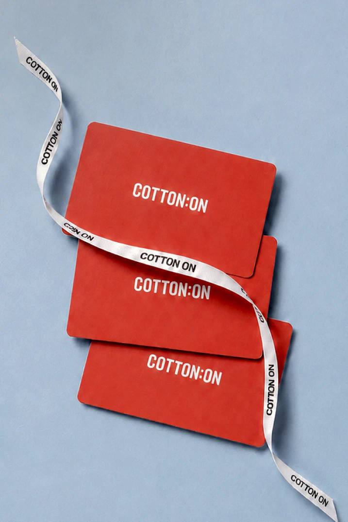 Two knit beanies stacked together—white on top, burgundy below—wrapped with white "COTTON ON" branded ribbon against a blue background